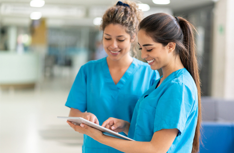 two medical assisting students in scrub attire standing close together and smiling while reviewing clinical documents