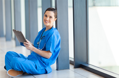An accelerated bachelor of science in nursing program student siting in hospital hallway, holding clinical documents and smiling. 